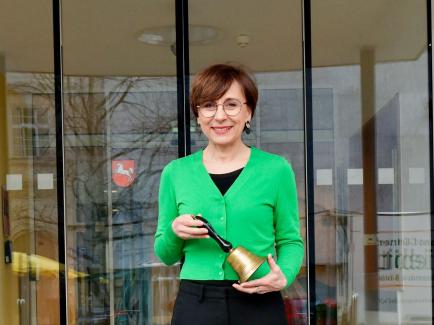 Braunhaarige Frau mit Brille bis zur Hüfte sichtbar in grünem Cardigan schaut lächelnd in die Kamera und hält eine Glocke in der Hand.