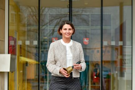 Braunhaarige Frau bis zur Hüfte sichtbar in hellem Jackett schaut lächelnd in die Kamera und hält eine Glocke in der Hand.