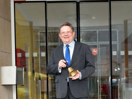 Braunhaariger Mann mit Brille bis zur Hüfte sichtbar in dunkelgrauem Jackett und mit blauer Krawatte schaut lächelnd in die Kamera und hält eine Glocke in der Hand.